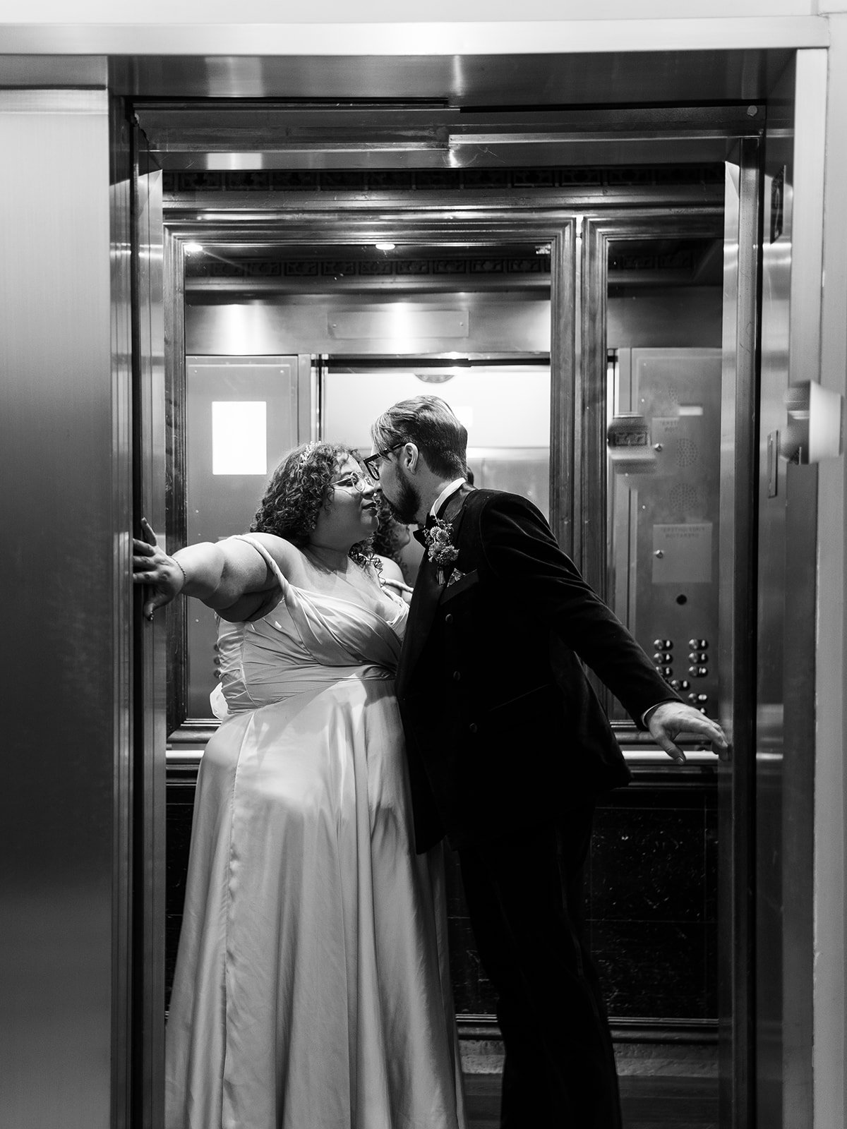 Black and white photo of bride and groom sharing a kiss inside a hotel elevator, each holding the doors open