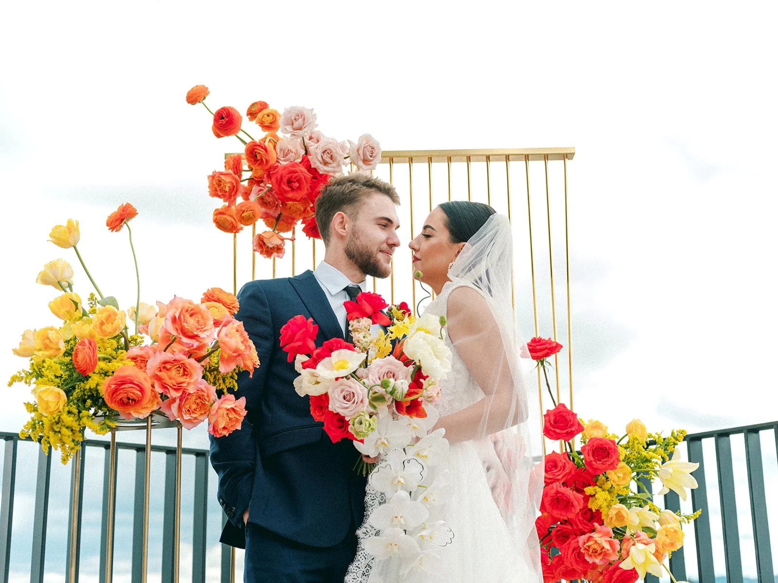 Bride and groom standing close together with vibrant floral installation on rooftop wedding backdrop