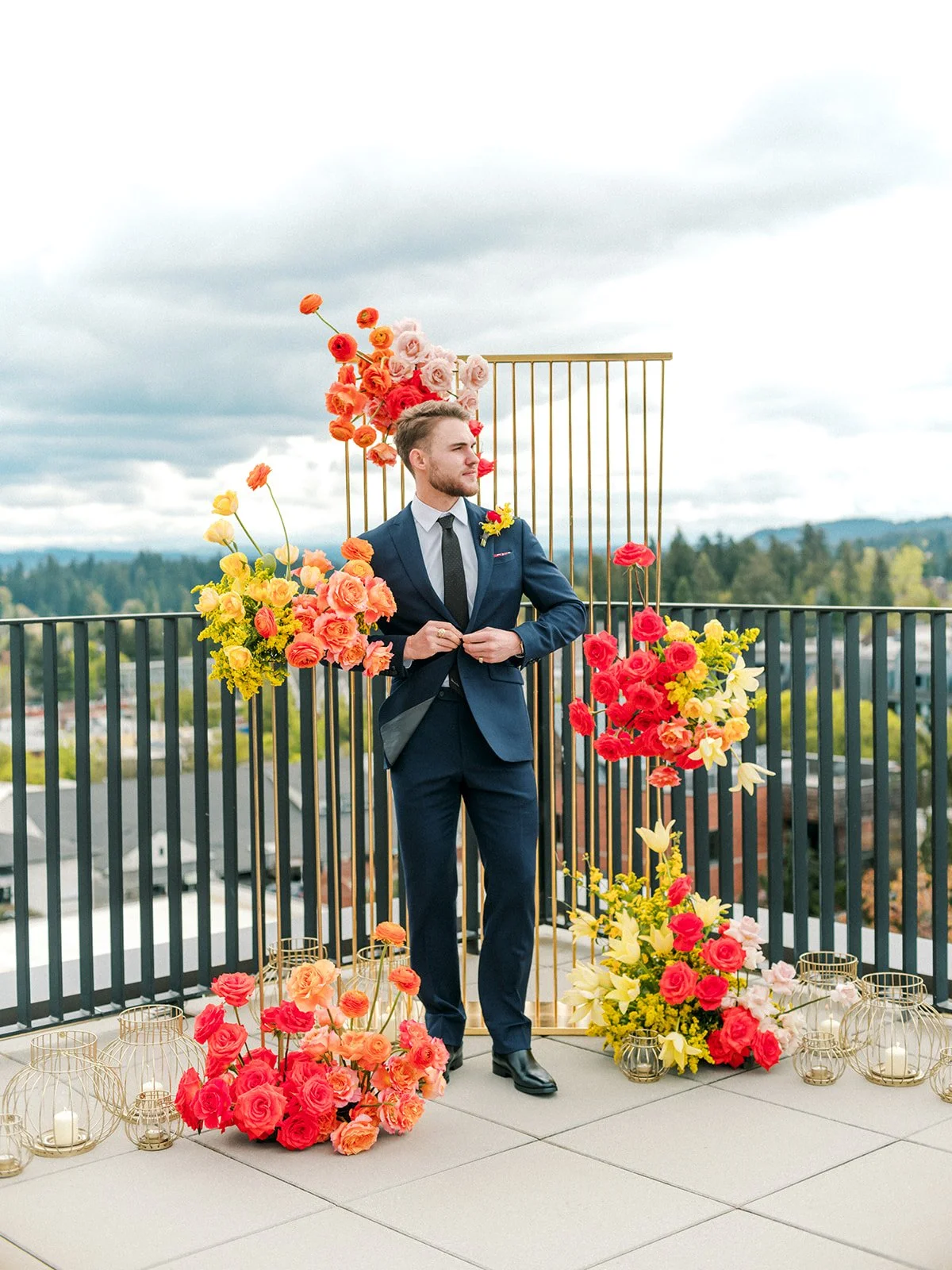 Groom adjusting suit jacket in front of bold floral ceremony backdrop on rooftop