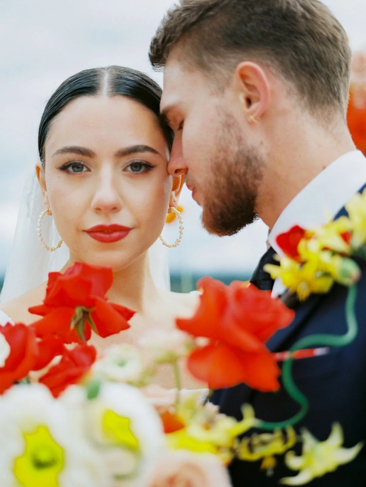 Close-up portrait of bride with bold lipstick and hoop earrings beside groom, framed by bright bouquet