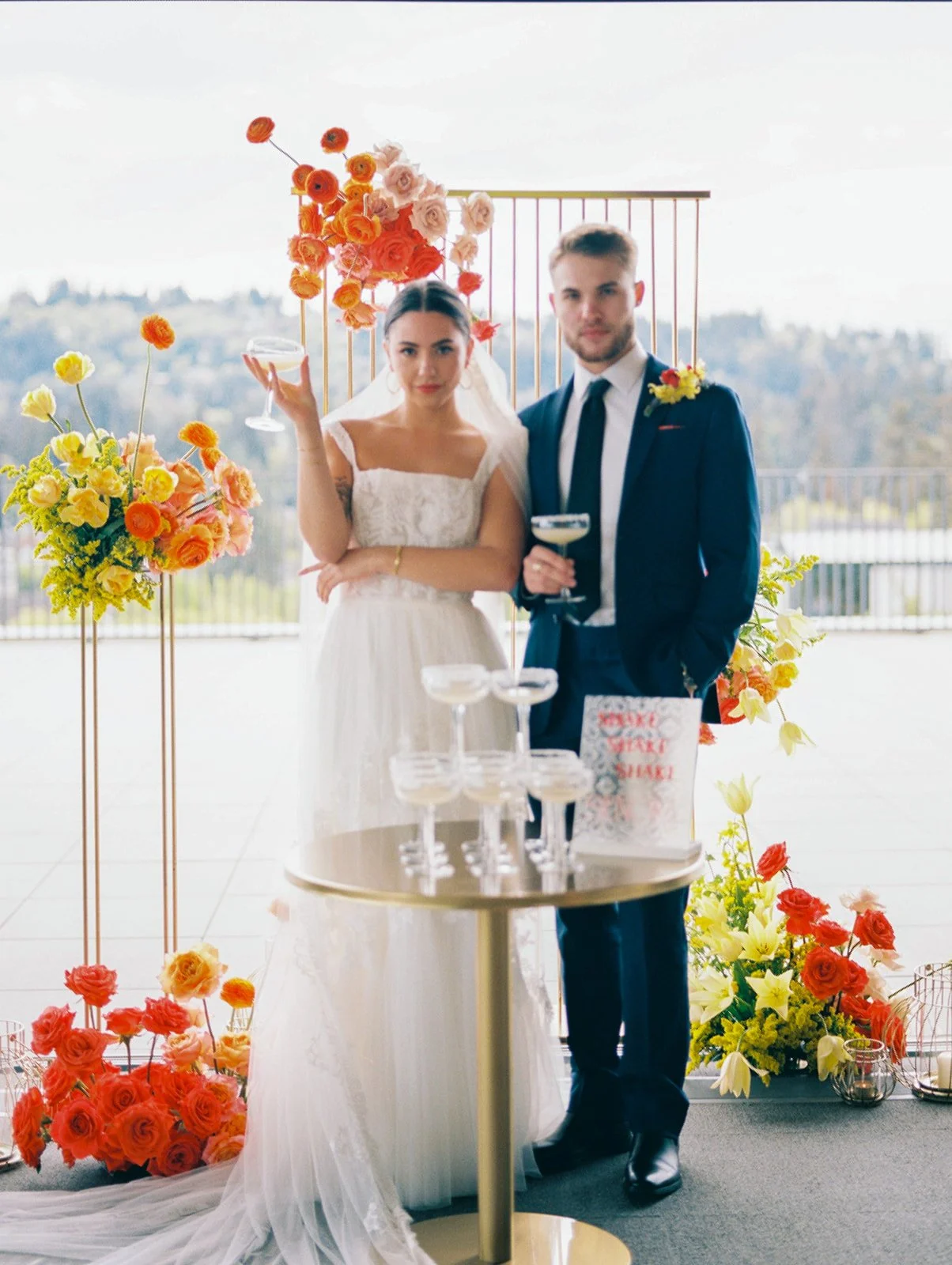 Bride and groom holding coupe glasses beside margarita tower and floral display at wedding reception