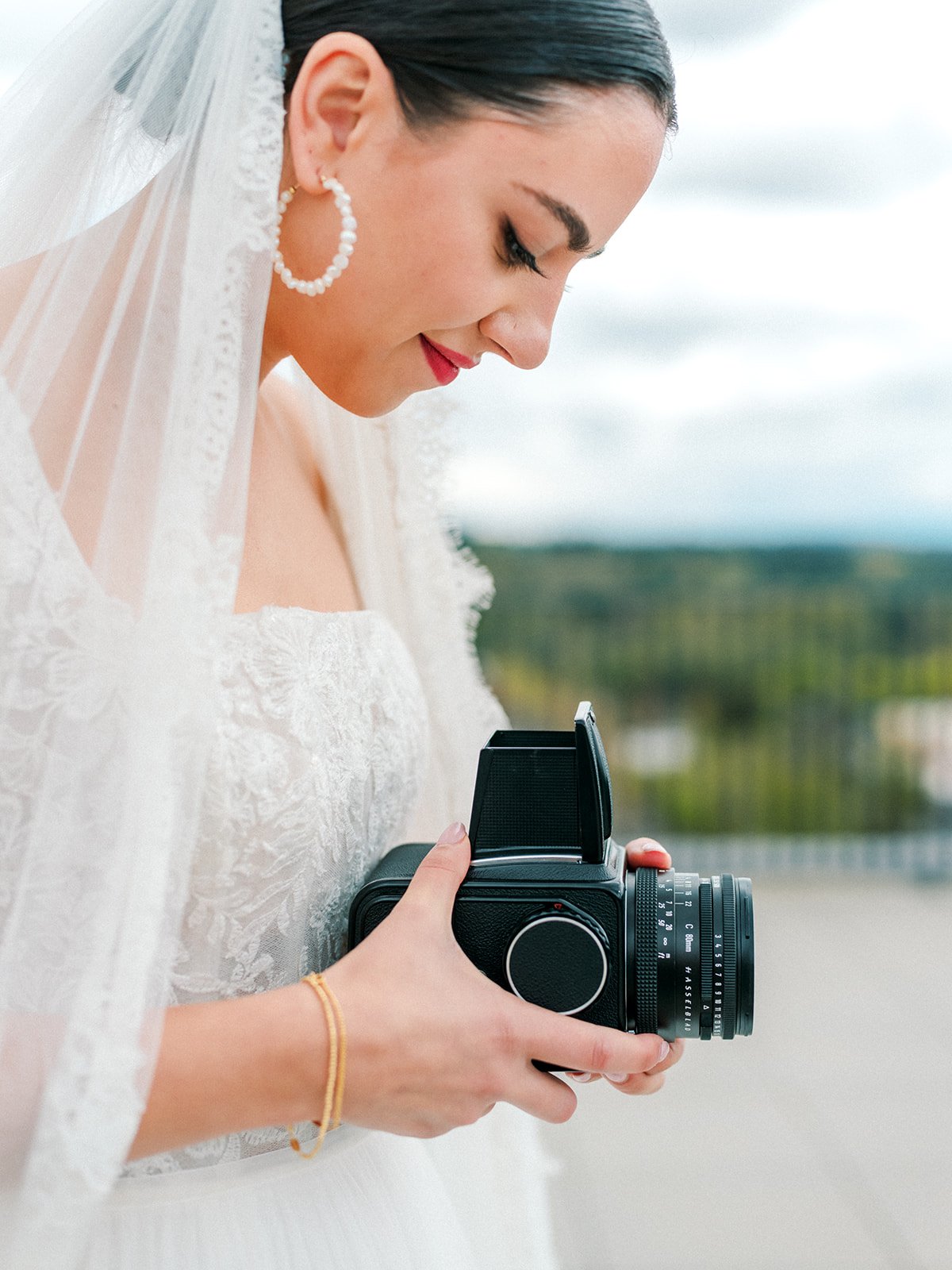 Bride in lace gown and veil holding vintage camera on rooftop terrace with soft natural light