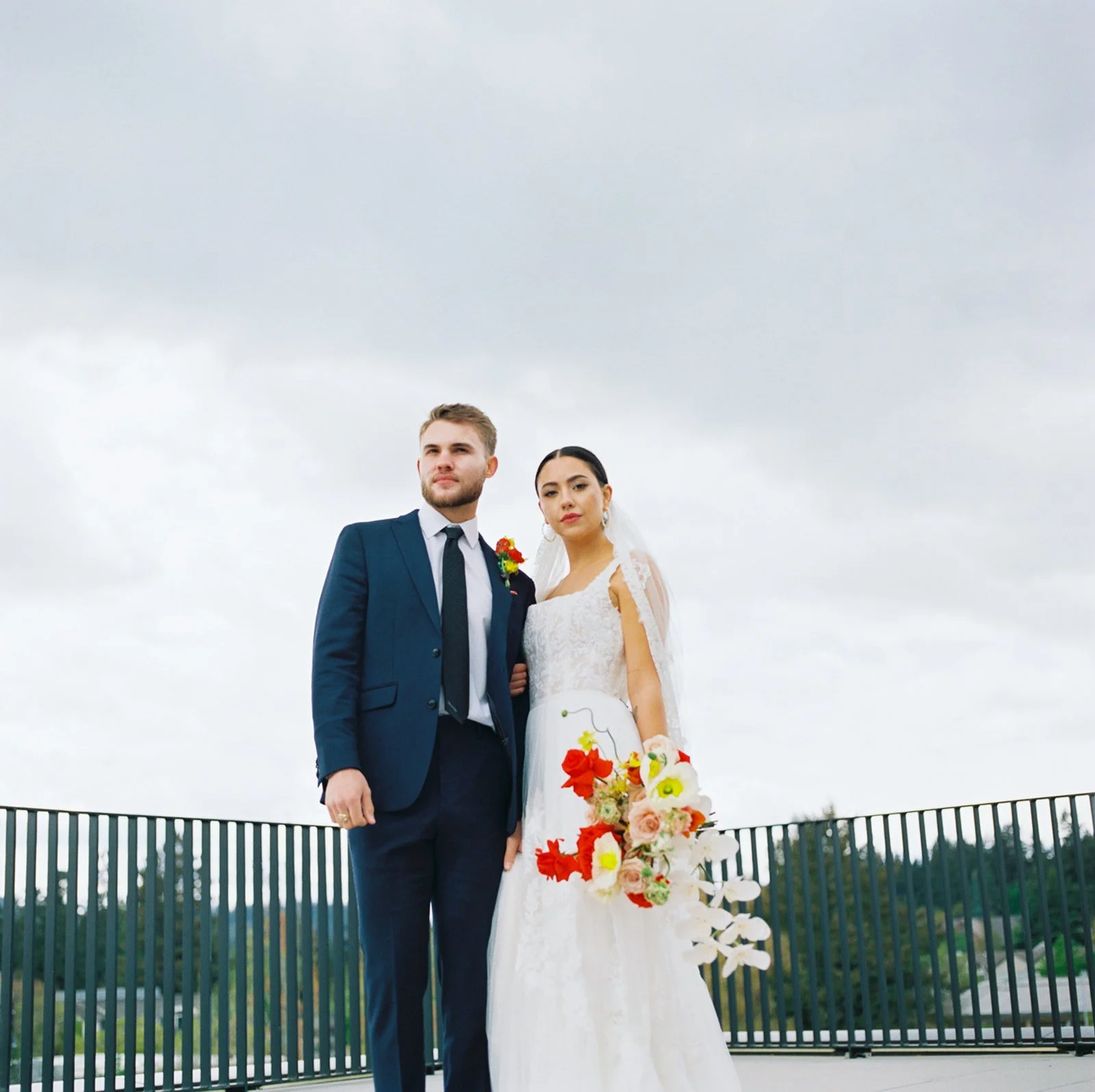 Bride and groom standing side by side on rooftop terrace holding bouquet against cloudy sky