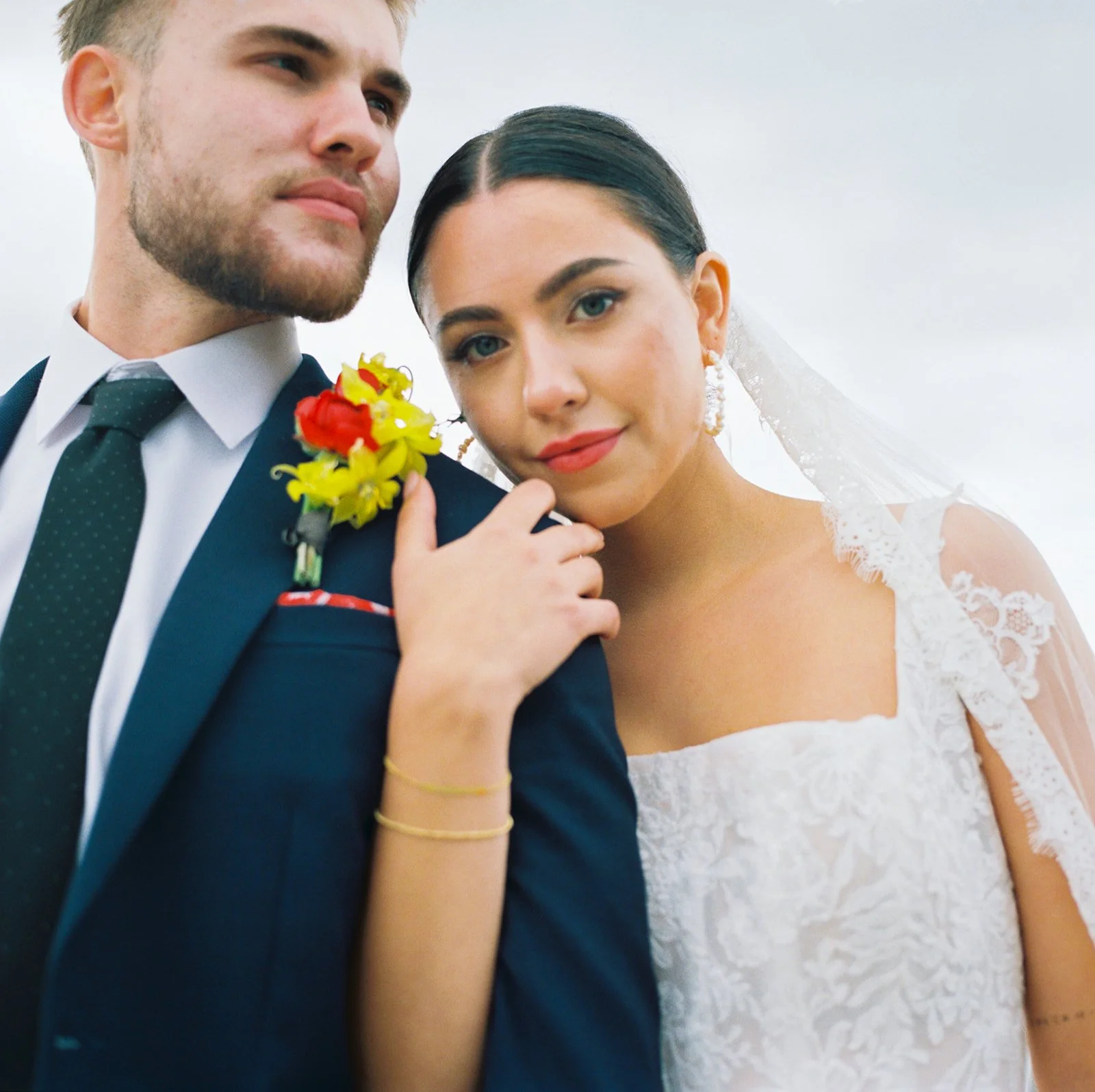 Close-up portrait of bride and groom with boutonniere and lace gown details against soft sky background