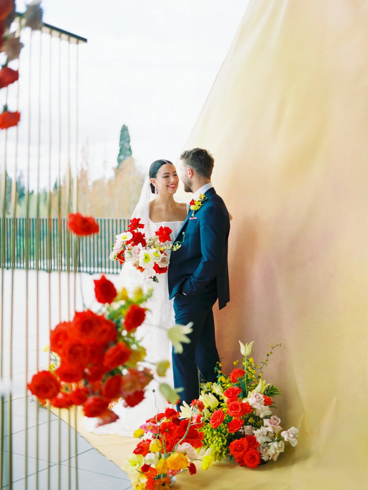 Bride and groom smiling together beside gold backdrop with vibrant floral arrangements at wedding