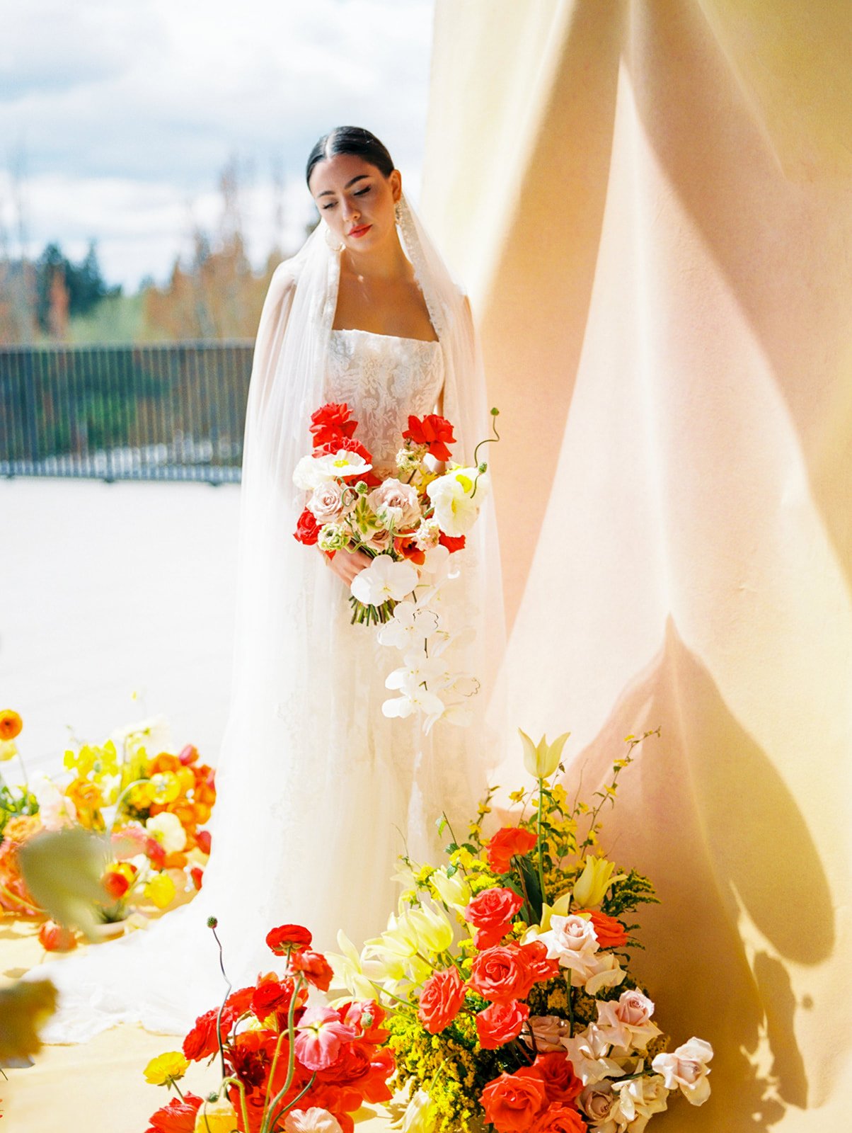 Bride holding colorful bouquet standing beside vibrant floral arrangements against soft golden wall