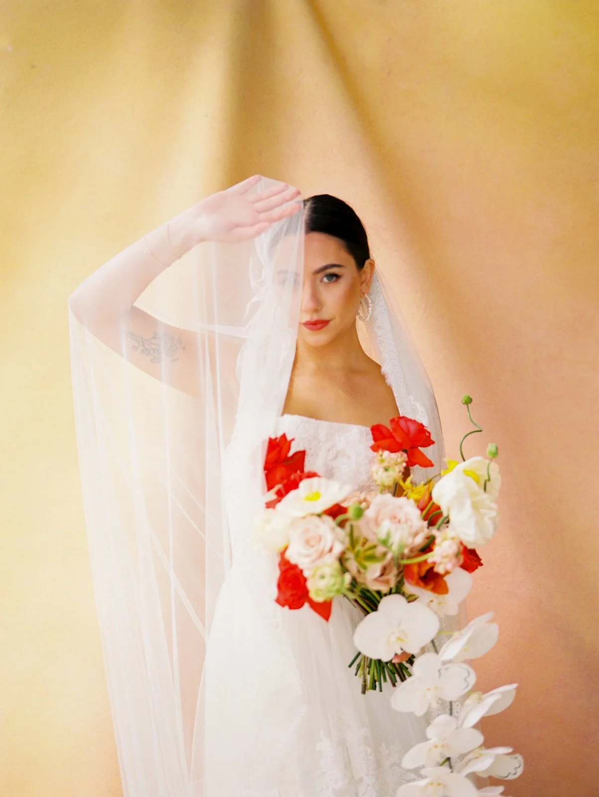 Bride holding veil over face with colorful bouquet against warm-toned backdrop