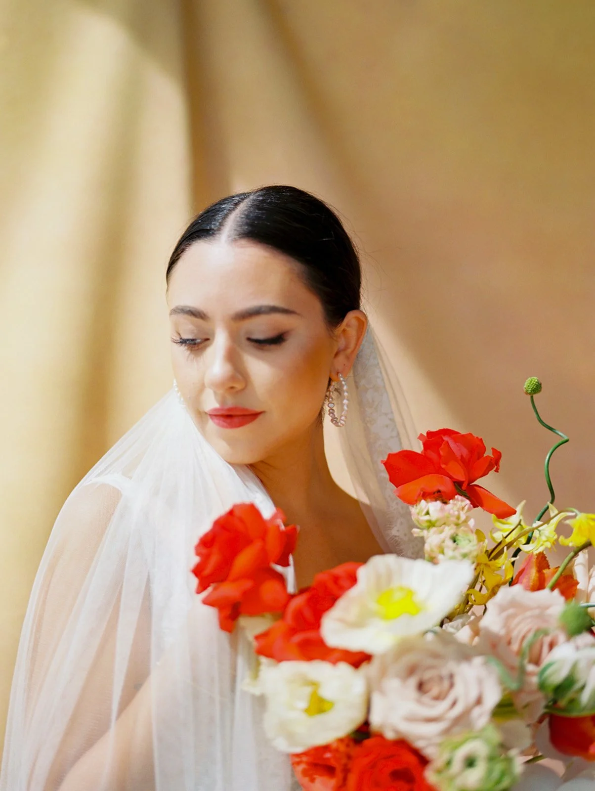 Bride in veil looking down at colorful bouquet with red, yellow, and blush flowers