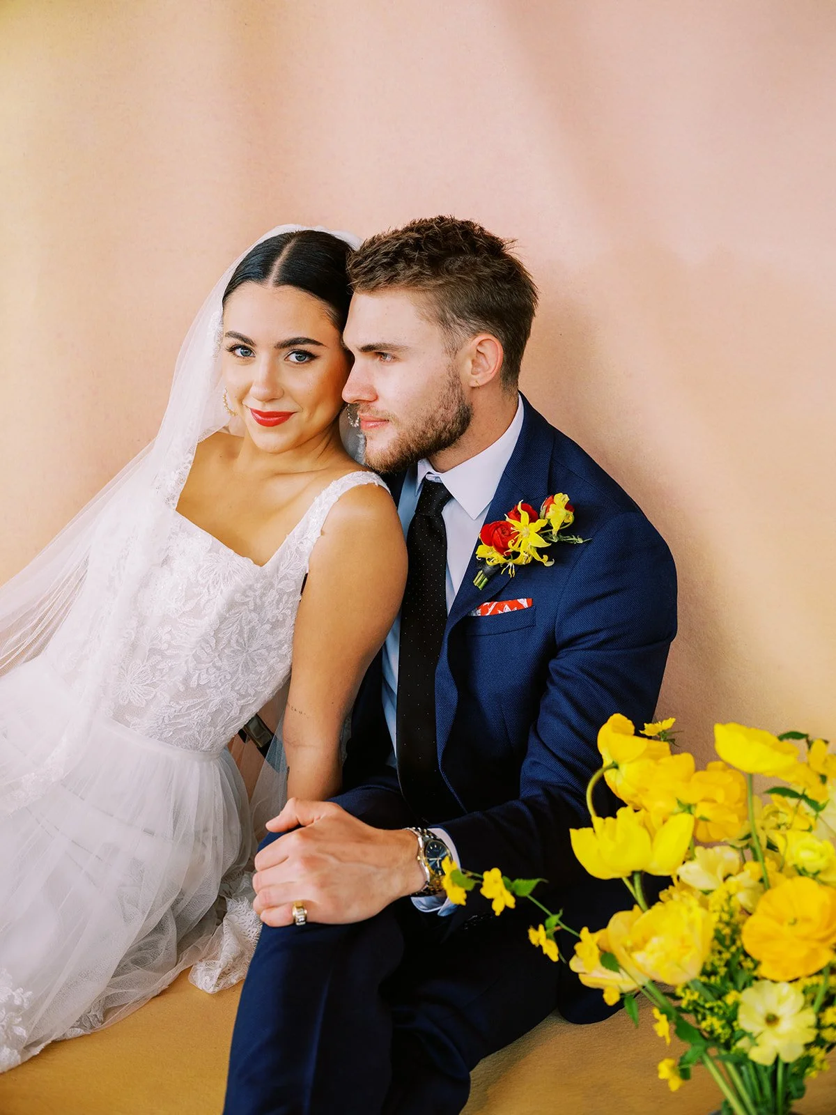Bride and groom seated together beside bright yellow flowers against warm-toned backdrop