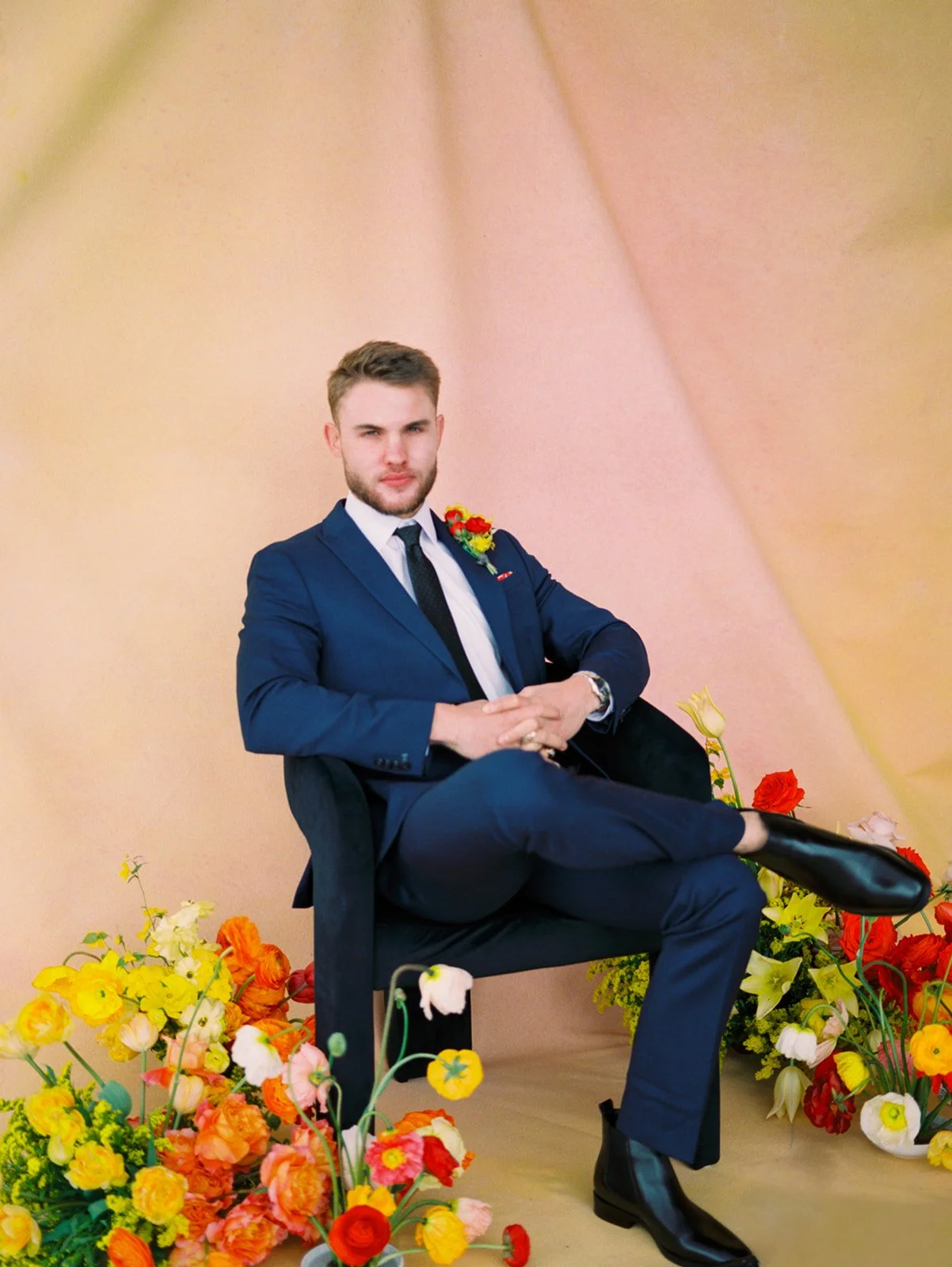 Groom seated in chair surrounded by colorful florals against warm-toned backdrop