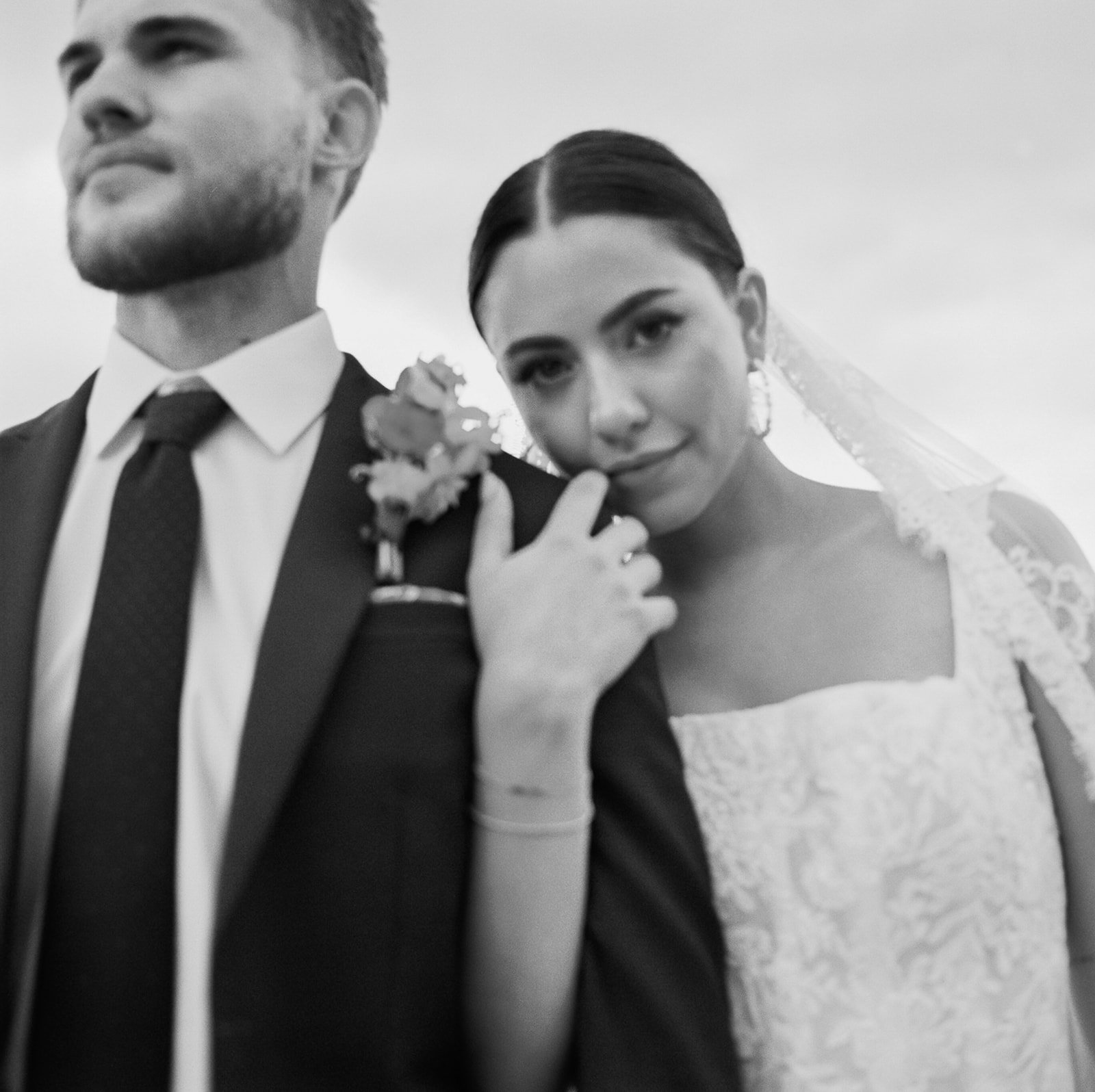 Black and white portrait of bride resting on groom’s shoulder, showing lace gown, veil, and boutonniere detail
