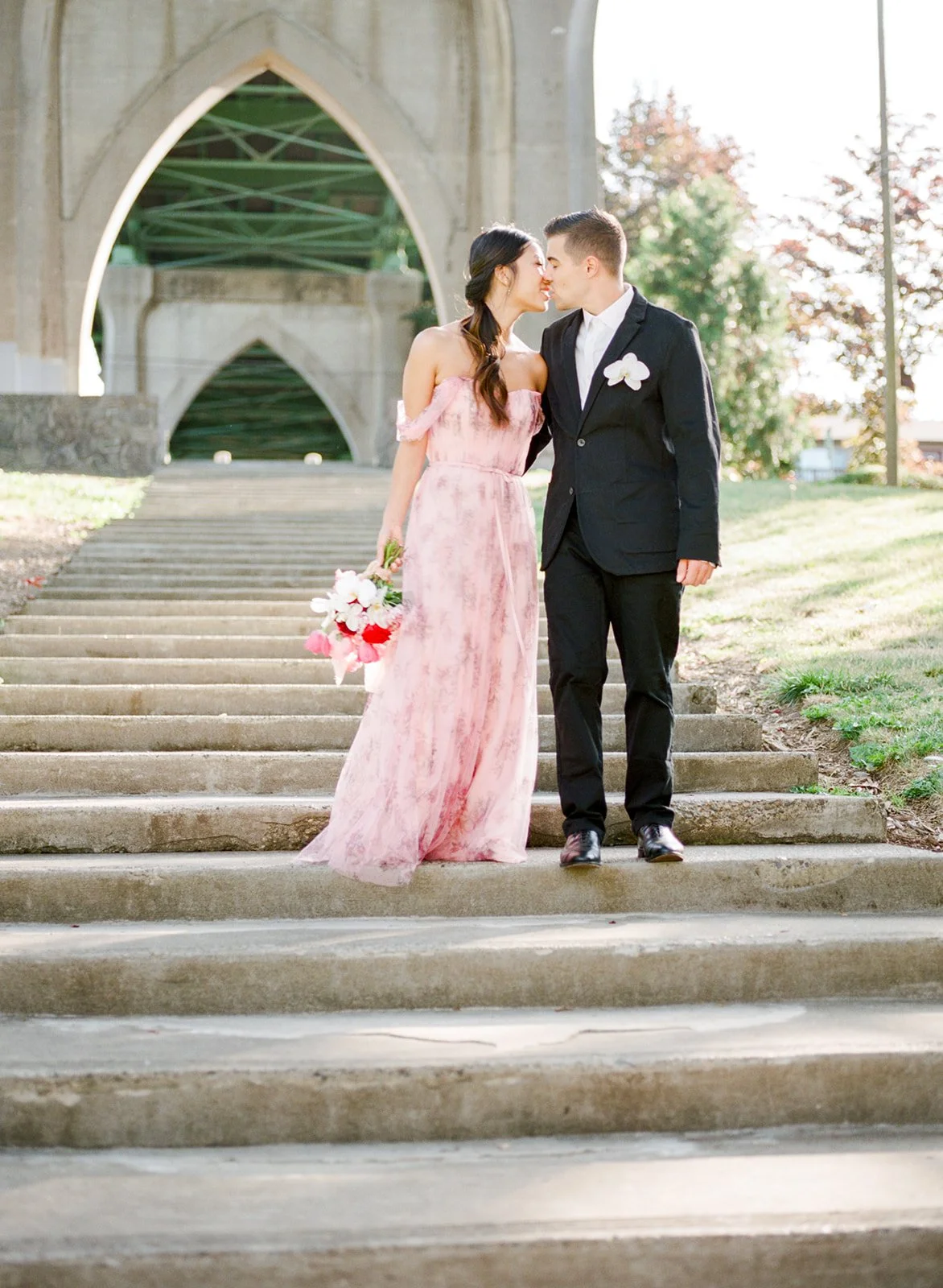 Couple in formal attire sharing a kiss on steps under Cathedral Park bridge arches.
