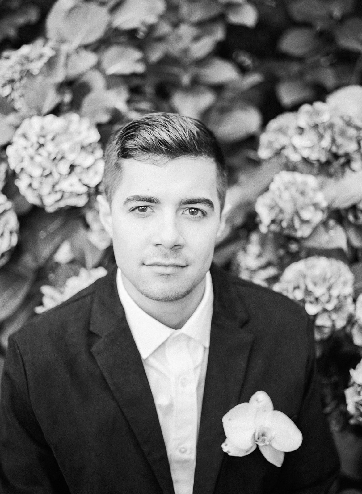 Groom in black suit portrait with boutonniere against leafy background at Cathedral Park.