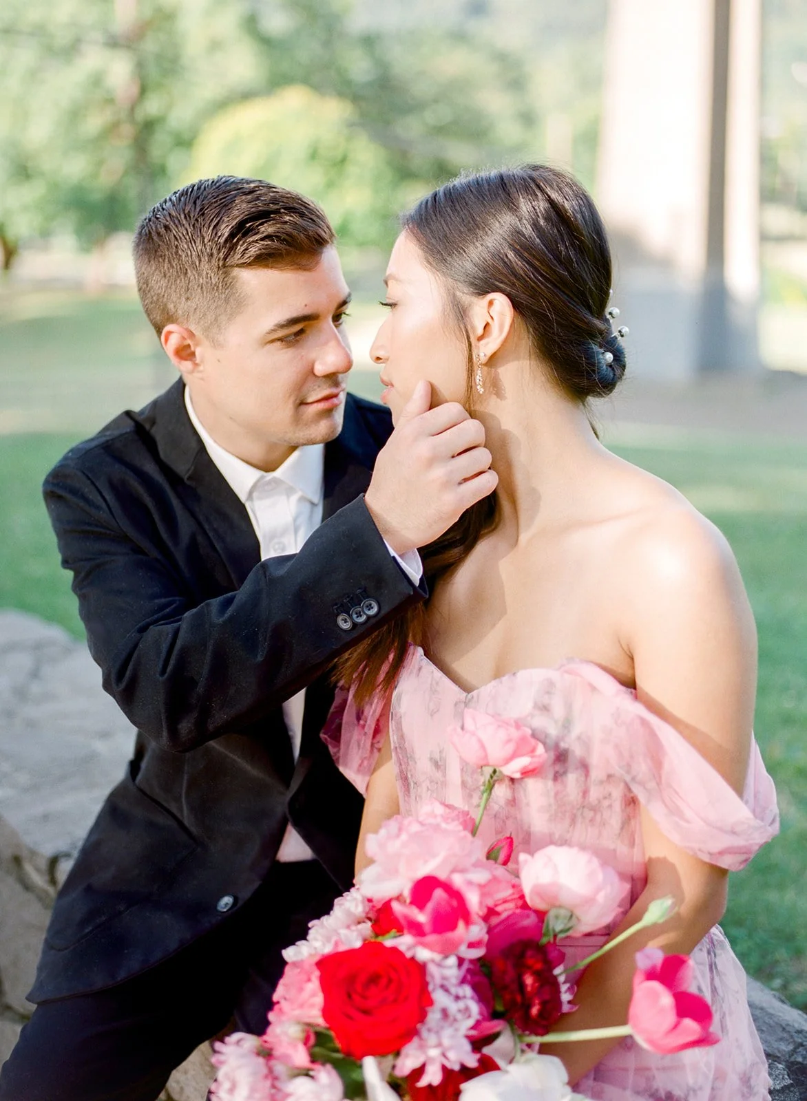 Groom gently touching bride’s chin as she holds pink and red bouquet in Cathedral Park.