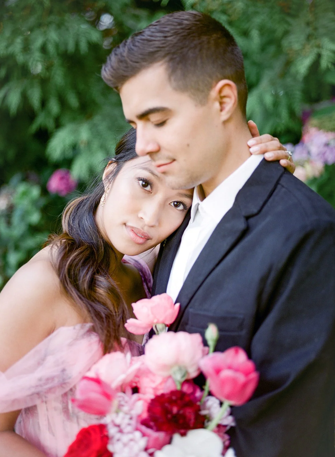 Close-up of couple embracing with bouquet, surrounded by lush greenery in Cathedral Park.