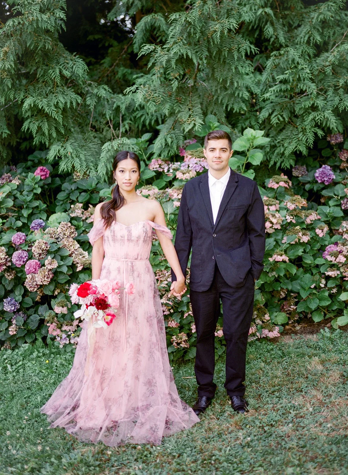 Couple standing hand in hand in front of hydrangea bushes at Cathedral Park.
