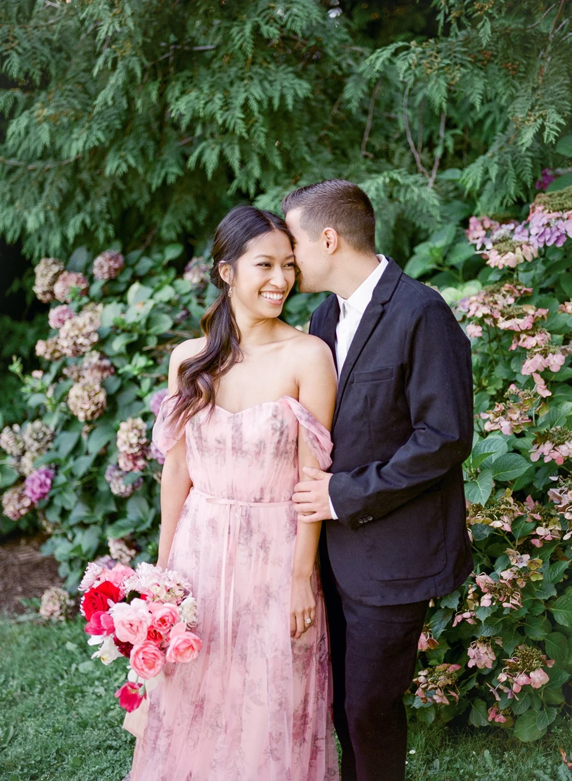 Groom leaning close as bride smiles holding bouquet among hydrangeas in Cathedral Park.