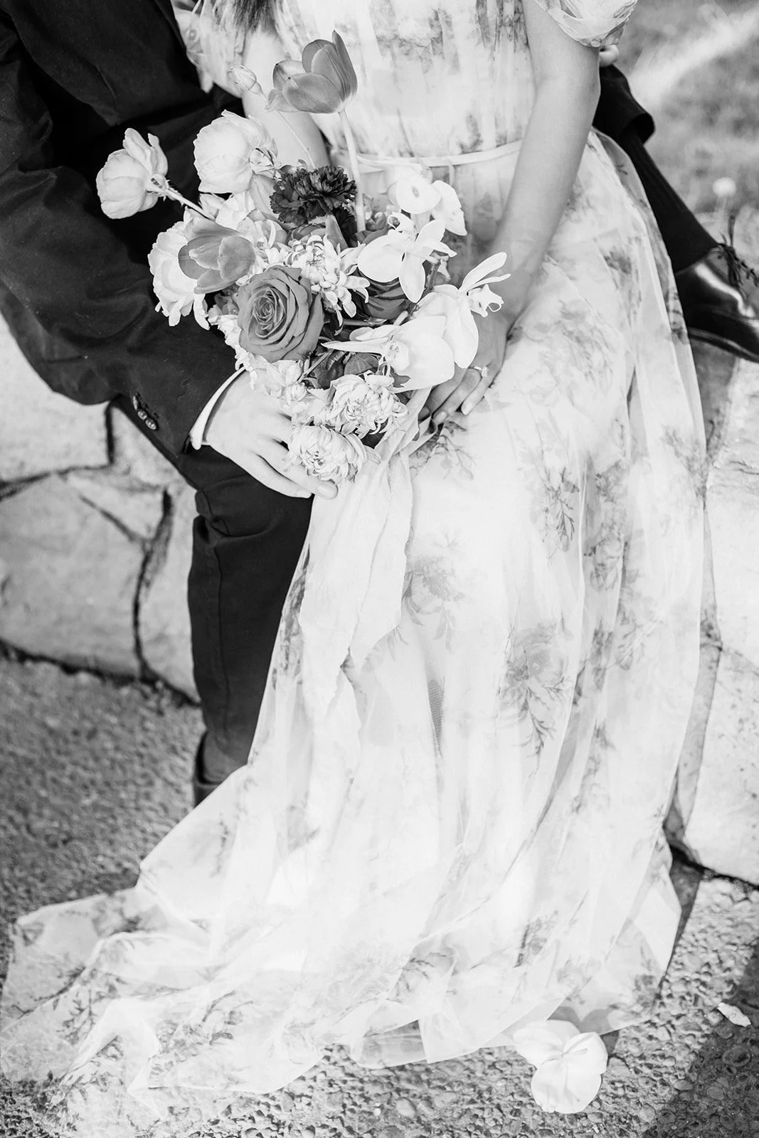 Close-up of couple holding a colorful bouquet, bride in floral gown and groom in suit against stone wall