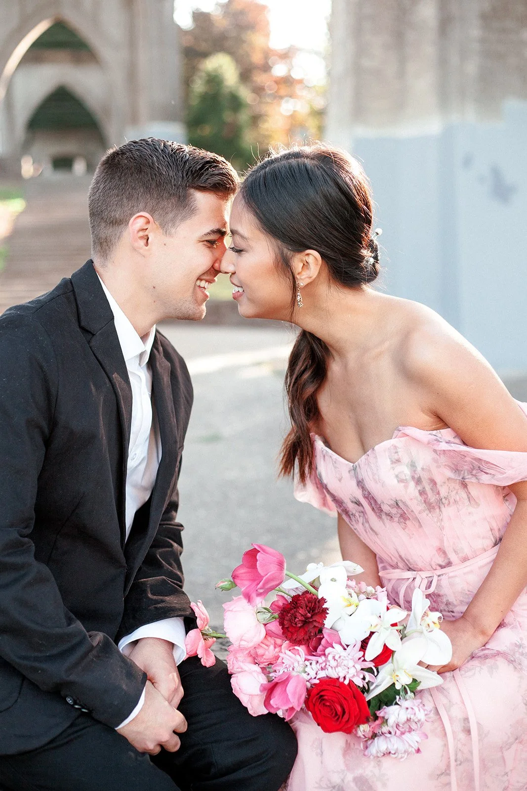 Couple sitting close, smiling with foreheads touching, bride holding pink and red bouquet in soft sunlight