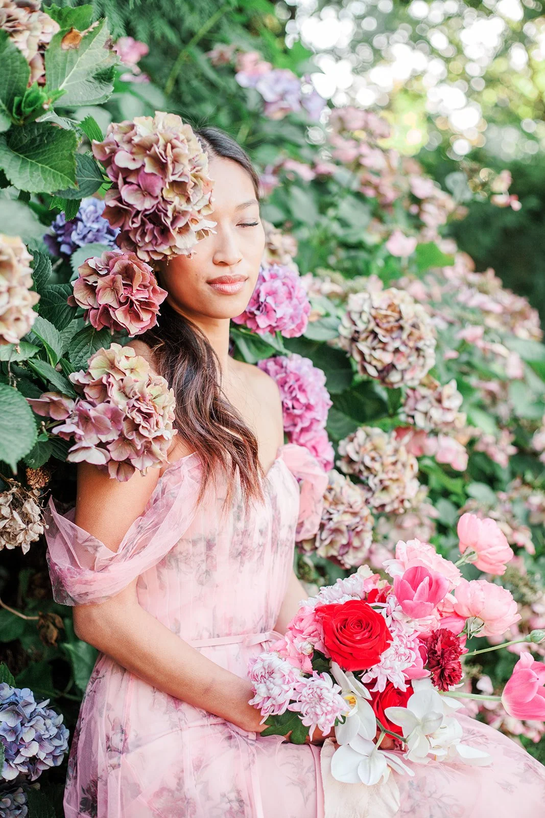 Bride in pink floral dress holding bouquet among blooming hydrangeas and greenery