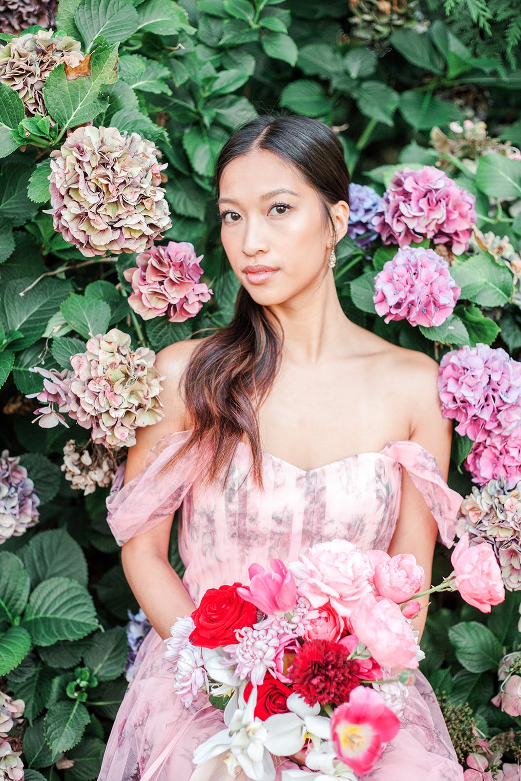 Portrait of bride in pink gown with bouquet, surrounded by lush green leaves and hydrangea flowers