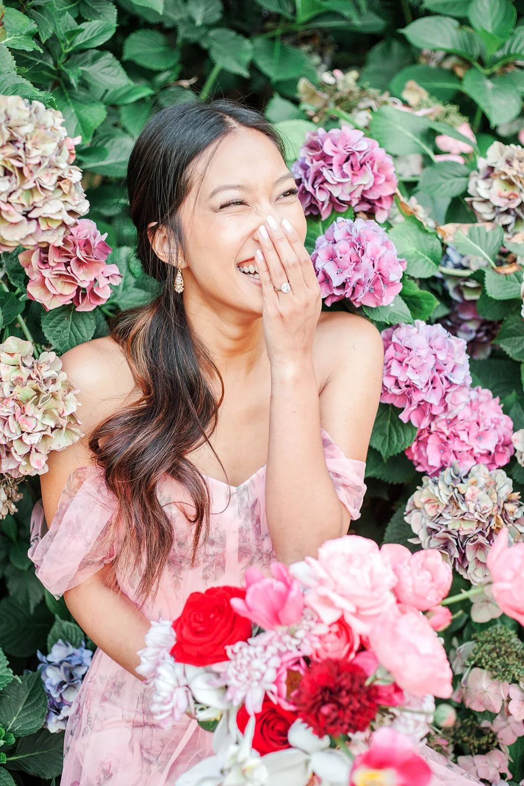 Bride laughing with hand near face, holding bouquet in front of blooming hydrangea bush