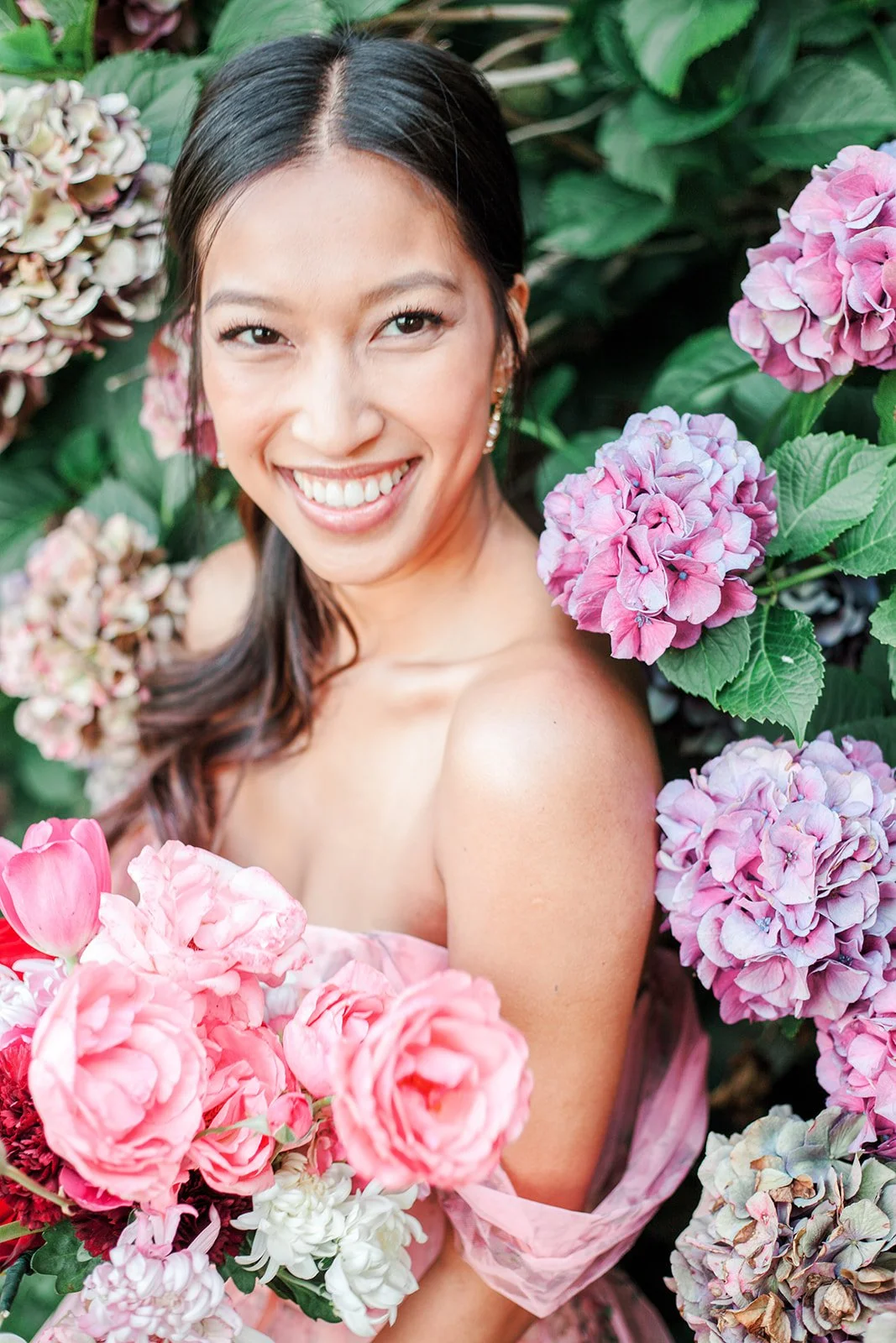 Close-up of smiling bride with bouquet, framed by pink and purple hydrangeas