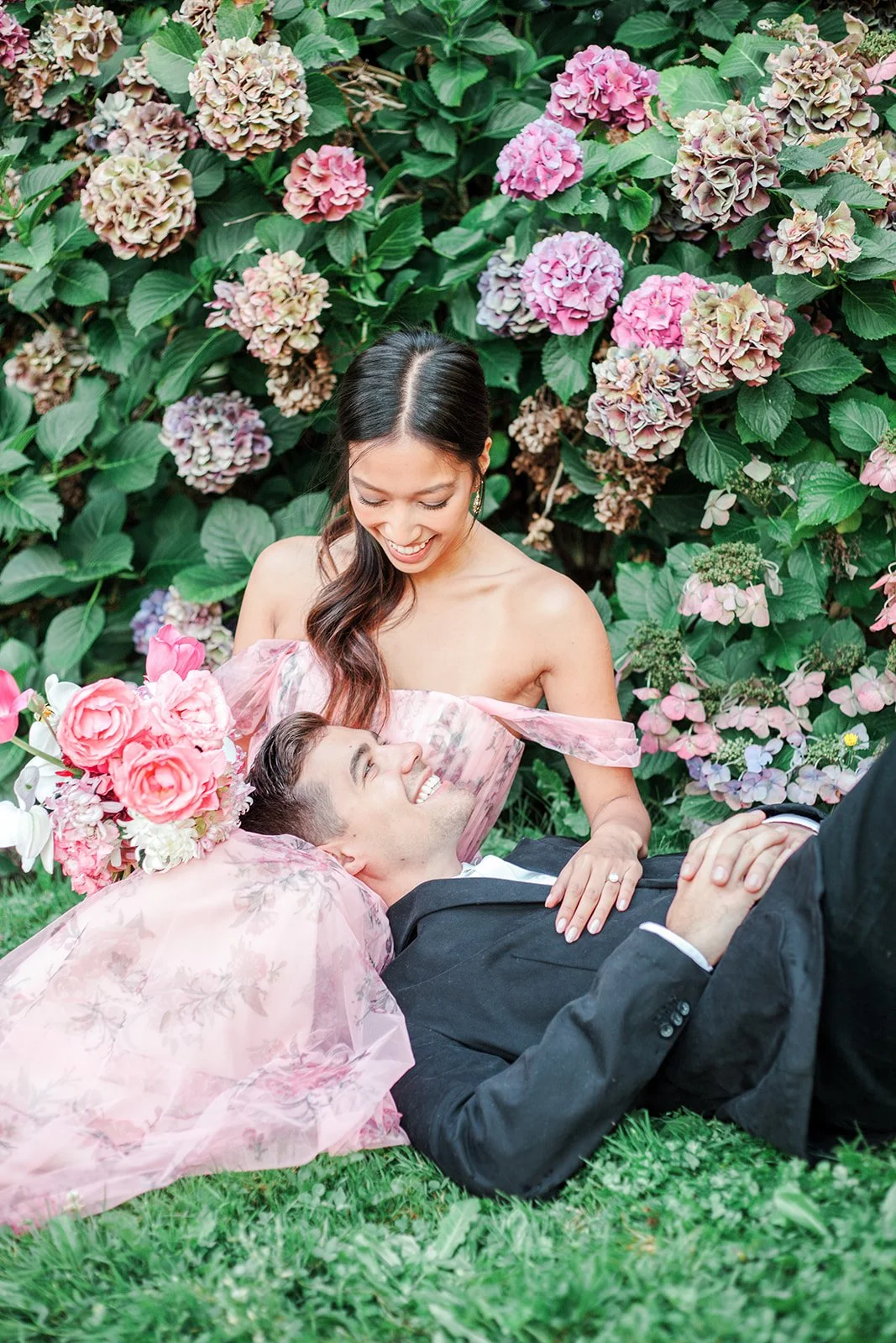 Couple lying on grass, bride leaning over groom while holding bouquet in garden setting