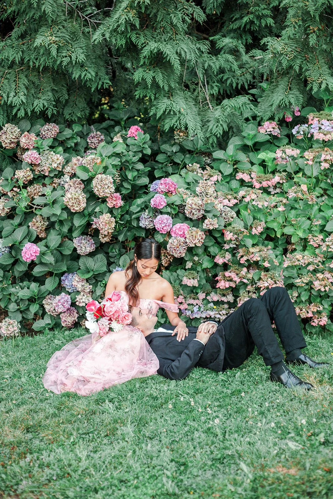 Wide shot of couple lying on grass near hydrangea bushes, bride looking down at groom
