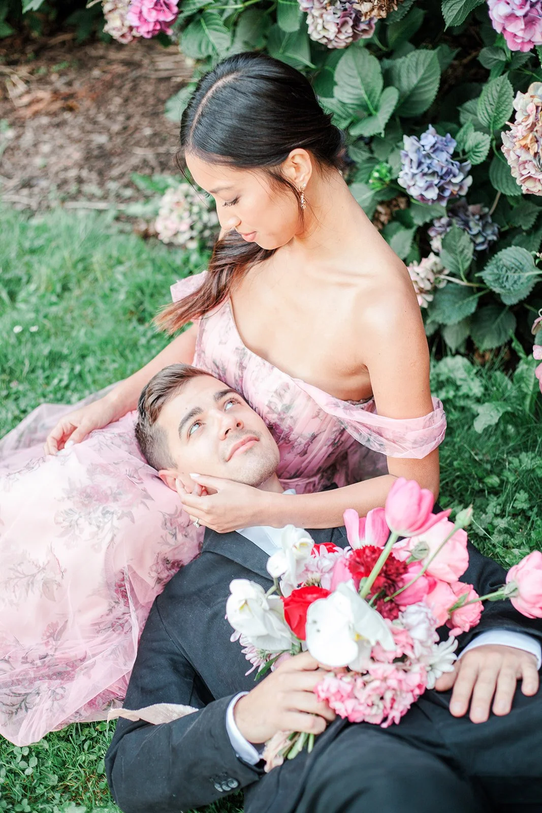 Close-up of bride cradling groom’s face as he lies on grass holding bouquet