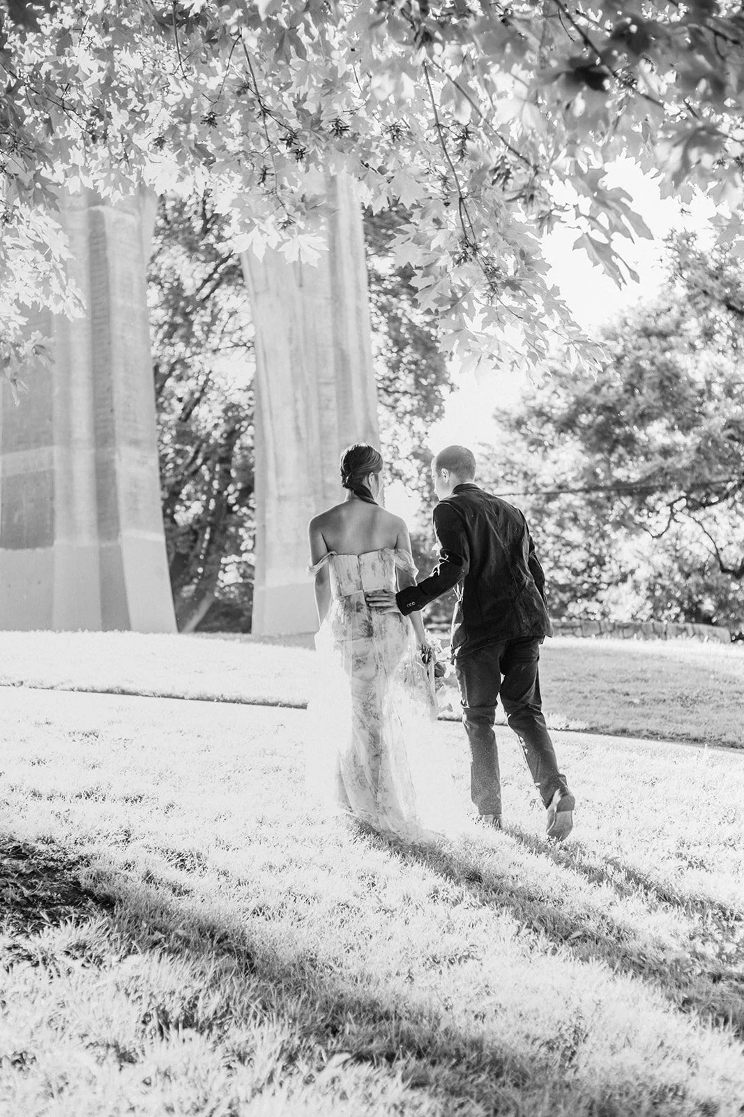 Black-and-white image of couple walking hand in hand beneath tall columns and trees