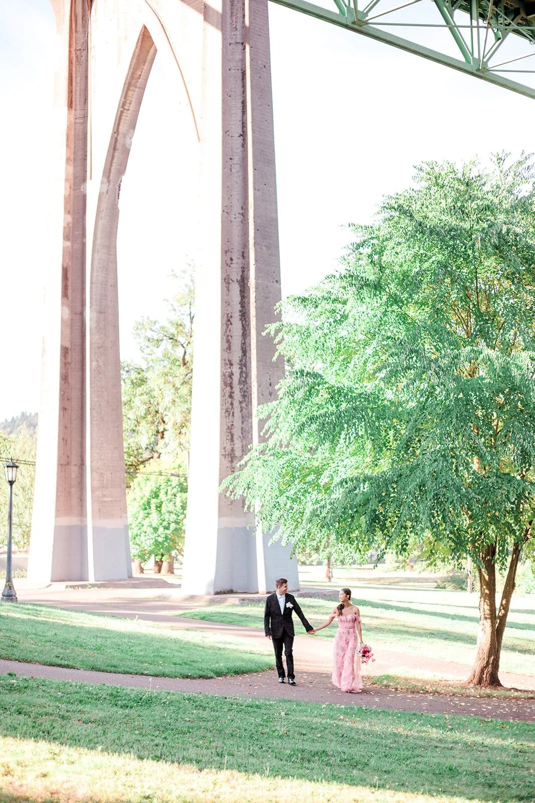 Wide shot of couple walking hand in hand under large bridge structure in park with trees