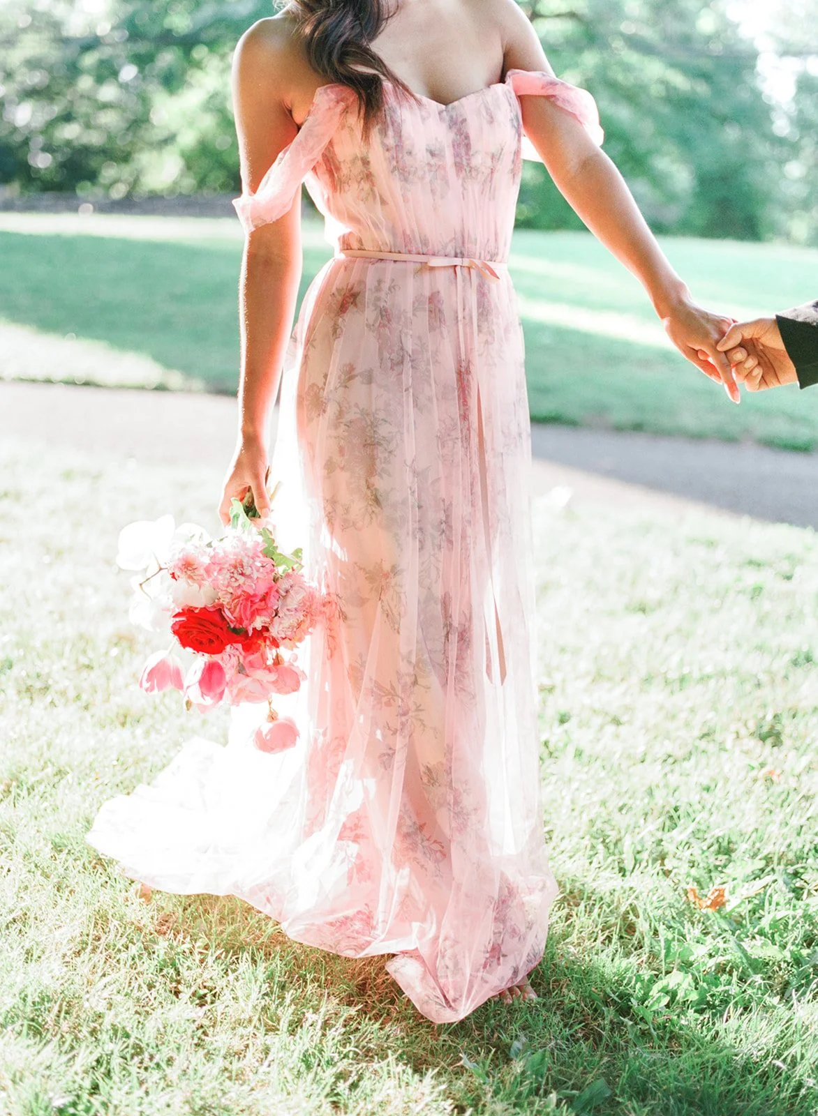 Woman in pink floral dress holding bouquet and leading partner by hand in sunlit Cathedral Park.