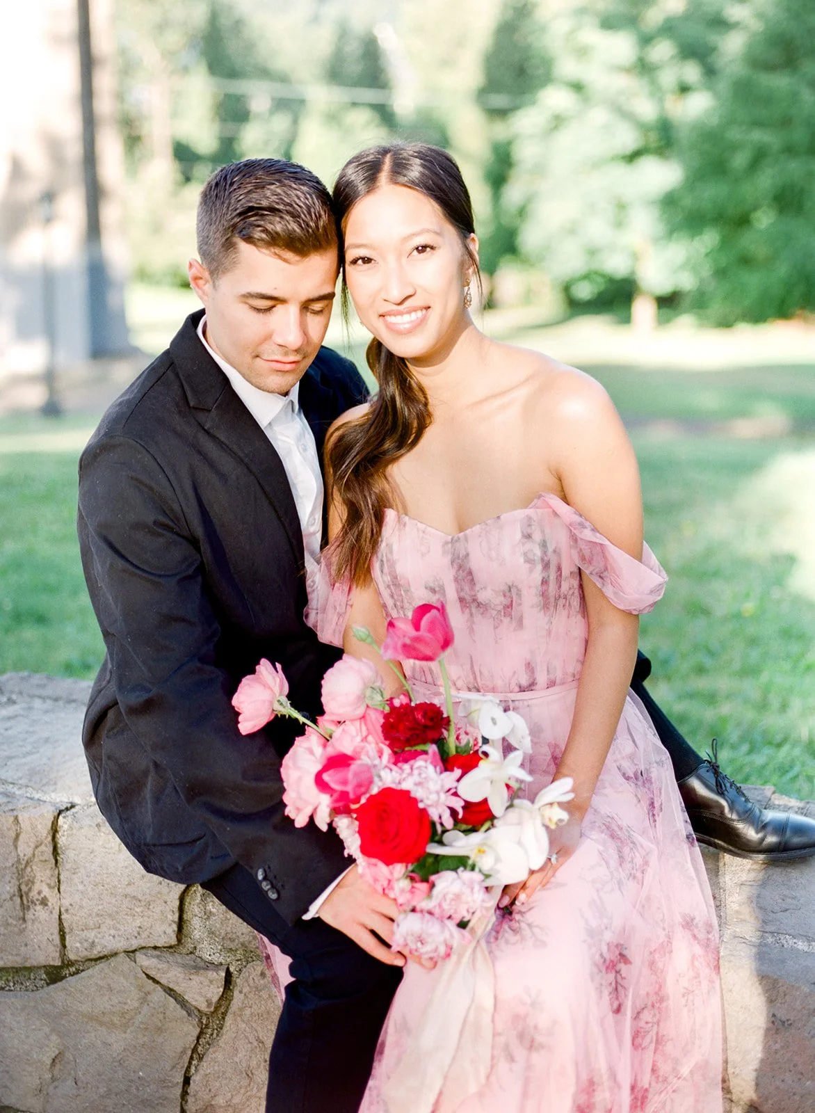 Couple seated on stone ledge with bouquet, soft light and greenery in Cathedral Park.
