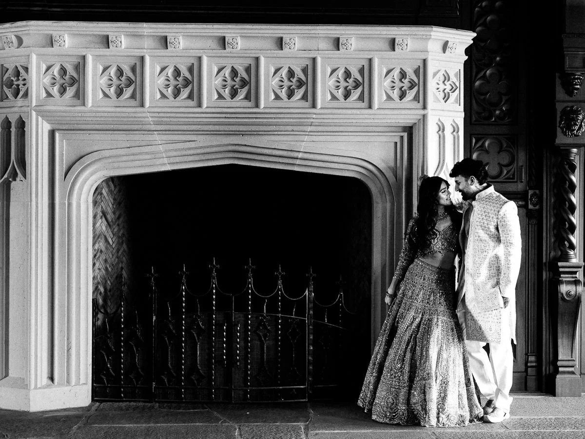 South Asian bride and groom standing inside grand stone fireplace in elegant pastel wedding outfits at Chateau de Lis.