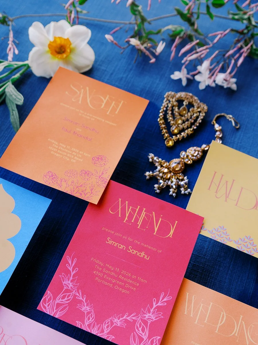 Bright pink and orange mehndi and haldi wedding invitations with gold jewelry and spring florals on a deep blue surface.