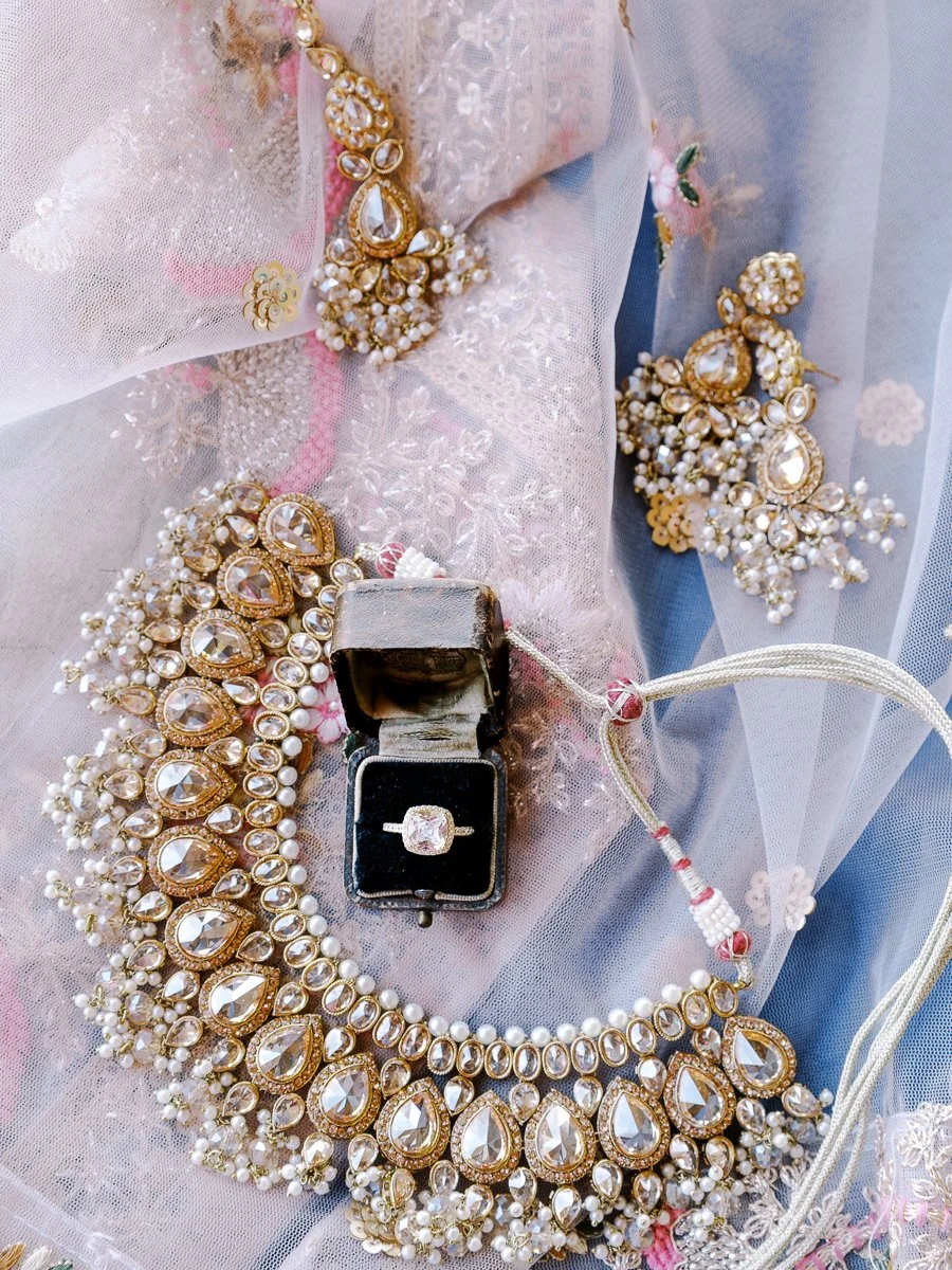 Gold bridal necklace, earrings, and diamond engagement ring arranged on embroidered blush fabric with pearl accents.