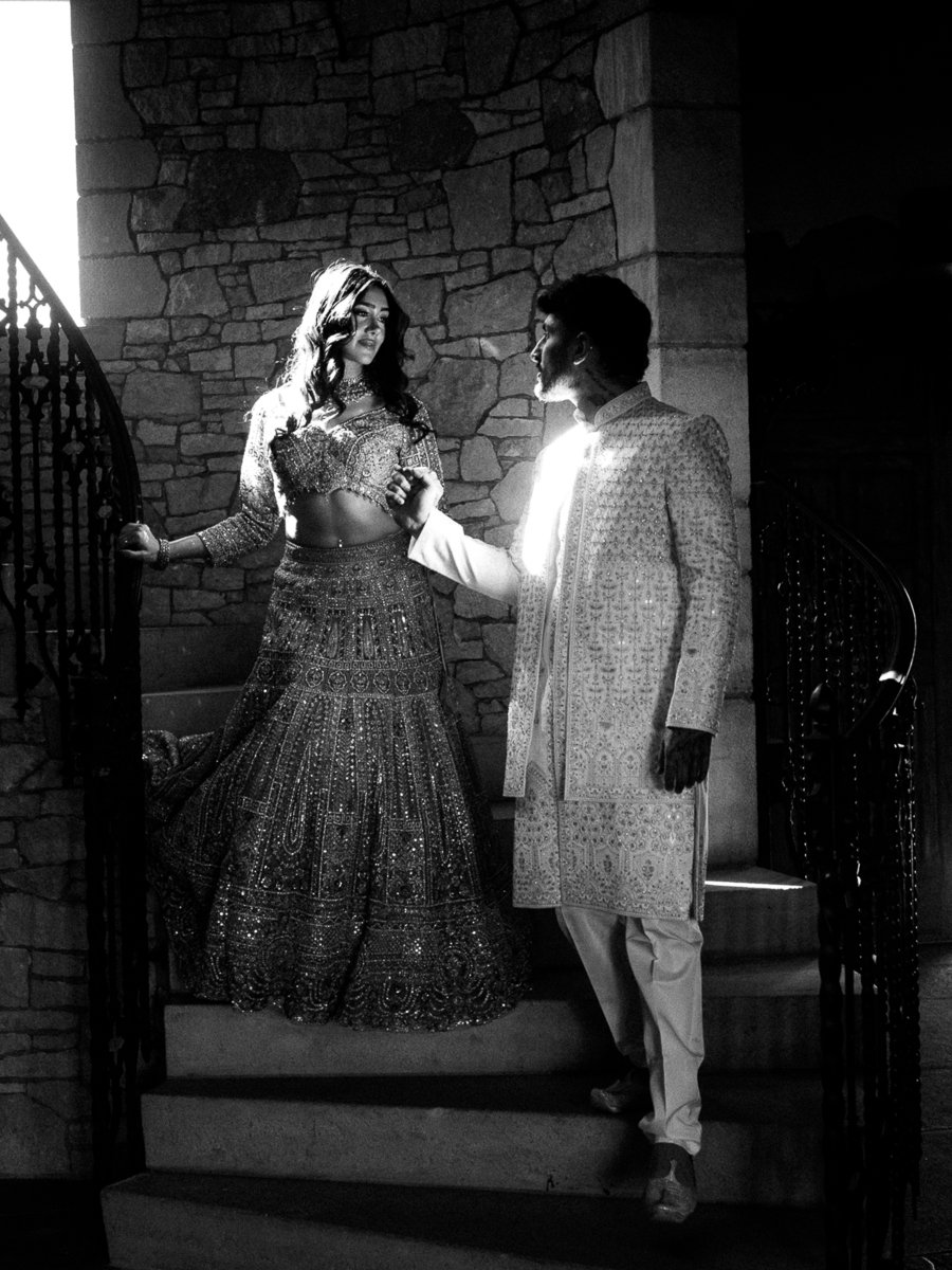 Black and white portrait of South Asian bride and groom holding hands on staircase with dramatic lighting and formal attire.