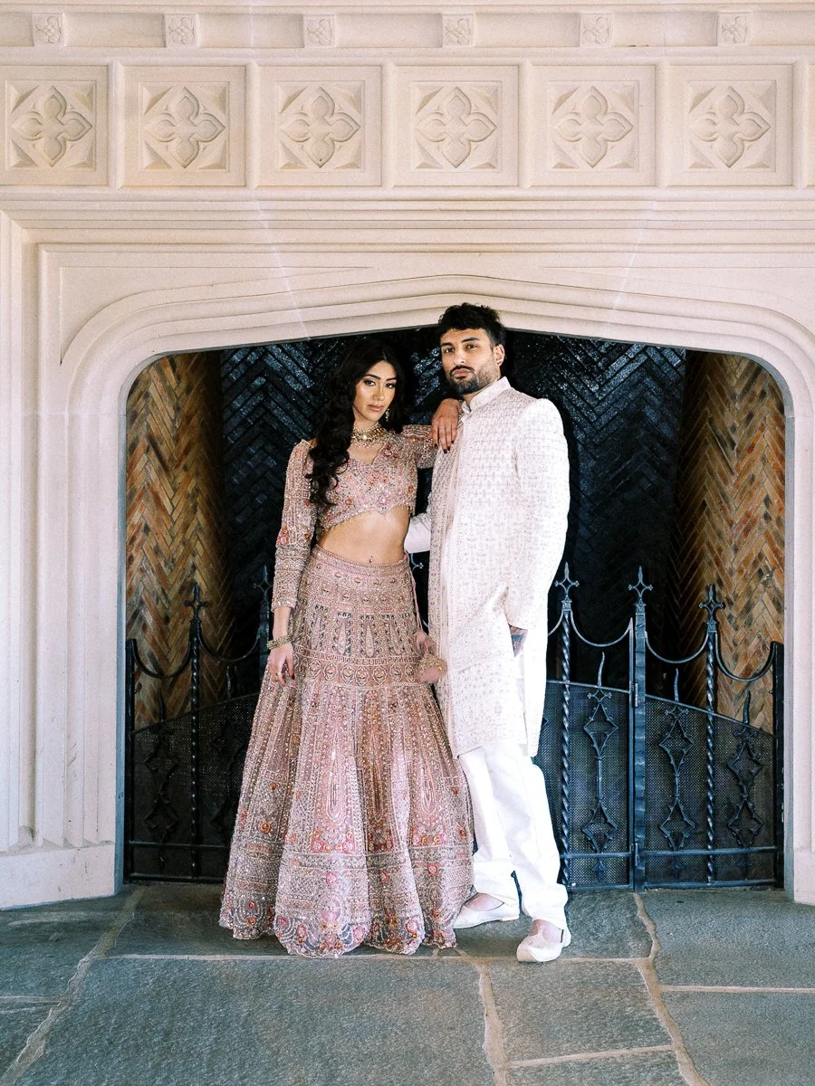 South Asian bride and groom standing inside grand stone fireplace in elegant pastel wedding outfits at Chateau de Lis.