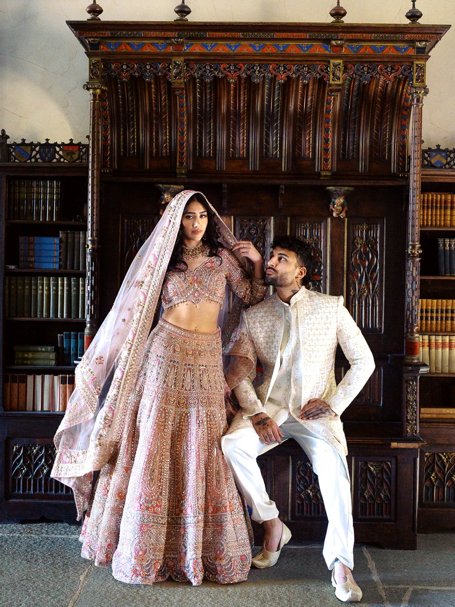 South Asian bride in embroidered blush lehenga and veil posing beside groom seated in ornate library at Chateau de Lis.