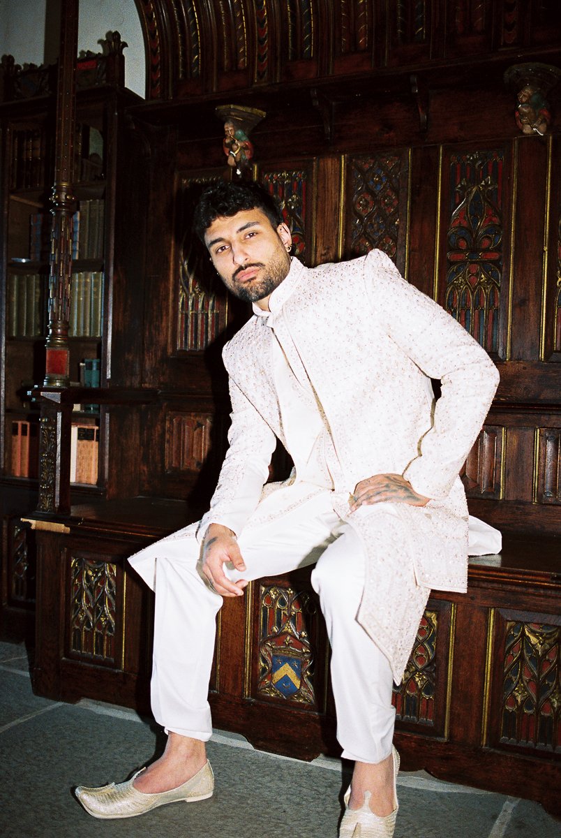 Groom portrait in ivory embroidered sherwani seated against dark wood architectural backdrop at luxury wedding venue.