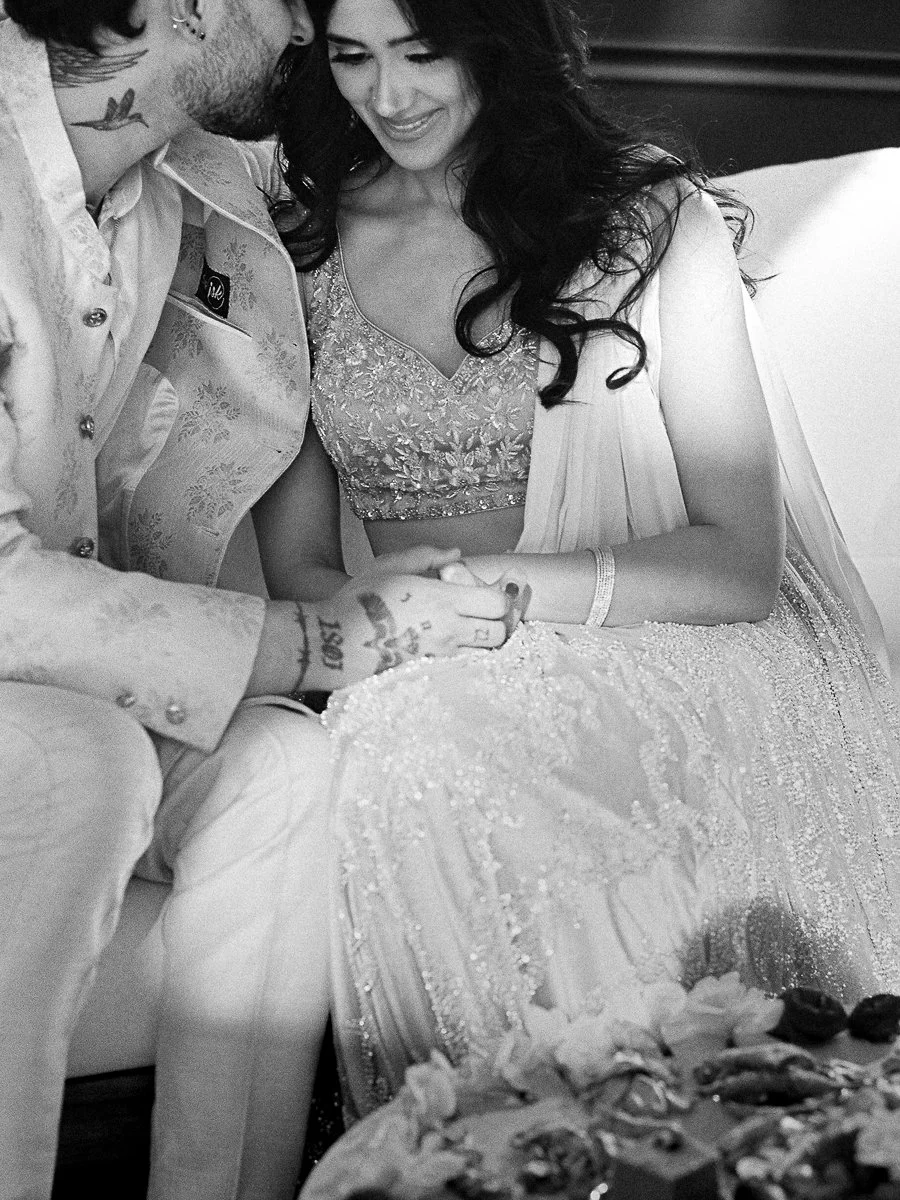 Black-and-white candid of couple seated together in pastel South Asian reception attire holding hands.