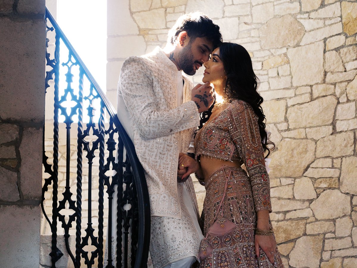 South Asian bride and groom posing on a stone staircase in embroidered pastel wedding outfits with dramatic backlighting.