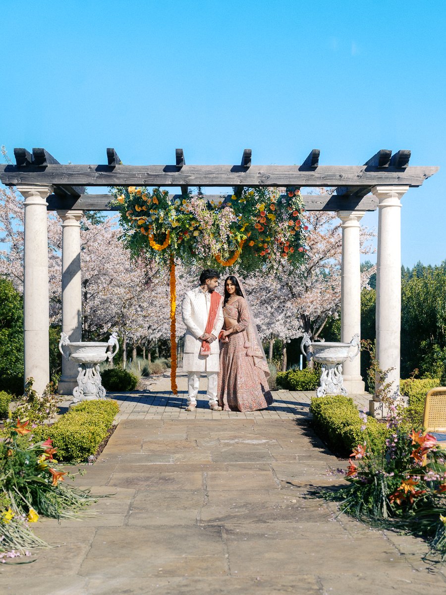 South Asian bride and groom standing beneath floral wedding mandap surrounded by blooming cherry blossoms at Chateau de Lis.