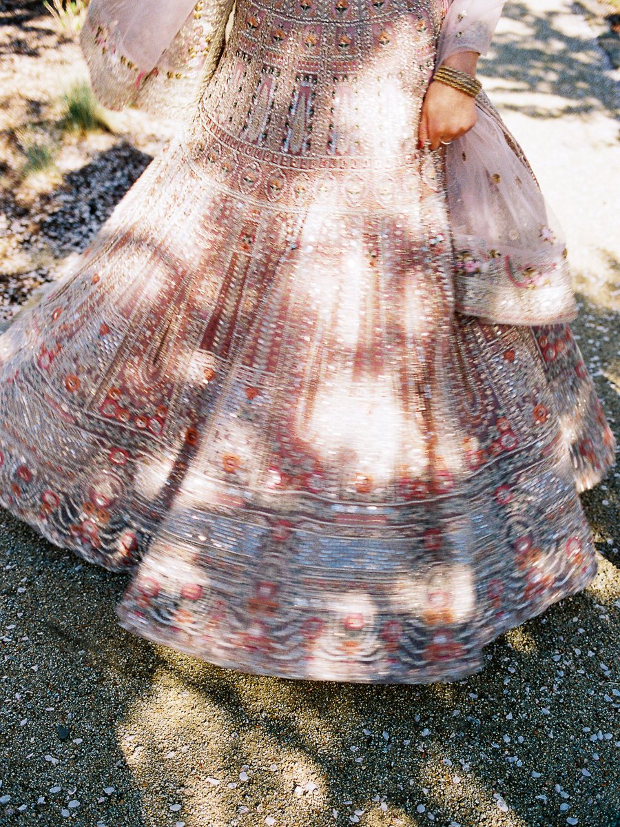 Motion detail of sparkling blush bridal lehenga twirling in sunlight during outdoor South Asian wedding portrait session.