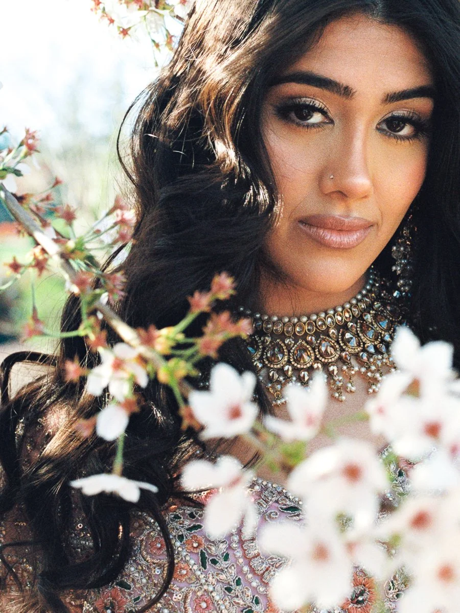 Bridal portrait framed by spring blossoms featuring ornate South Asian jewelry and embroidered pastel lehenga details.
