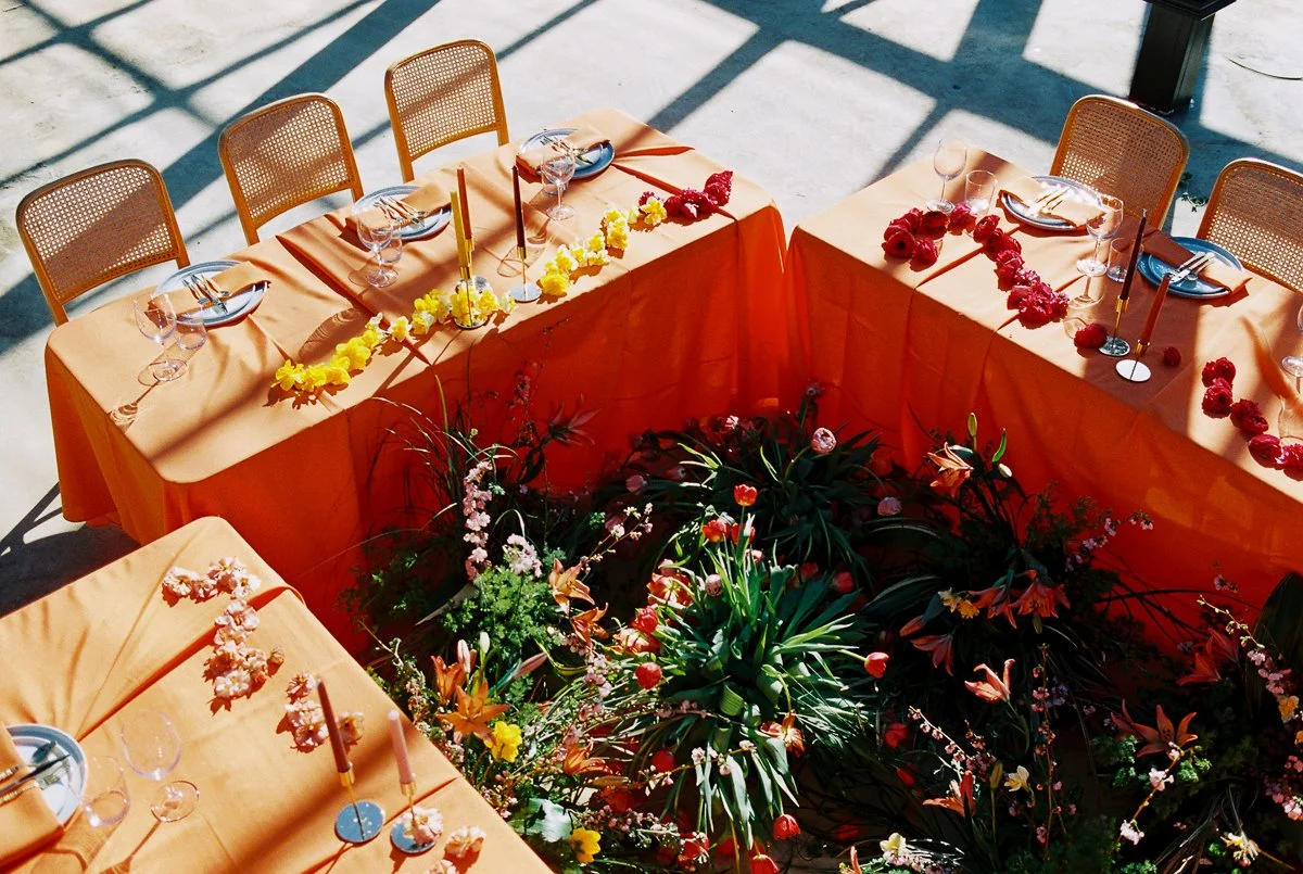 Colorful wedding reception tablescape with orange linens, floral centerpieces, and modern candle styling inside glass conservatory.