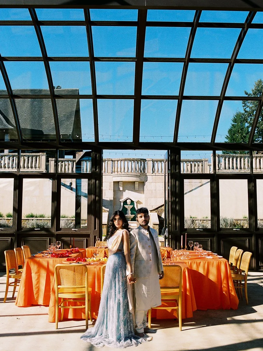 South Asian couple standing inside modern glass reception space with vibrant orange tablescape and dramatic natural light.