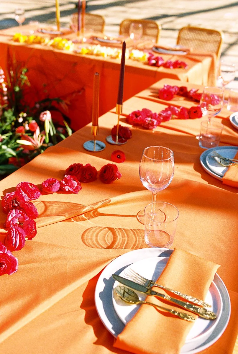 Bright wedding reception table setting with orange linens, floral garlands, gold flatware, and modern candle holders.