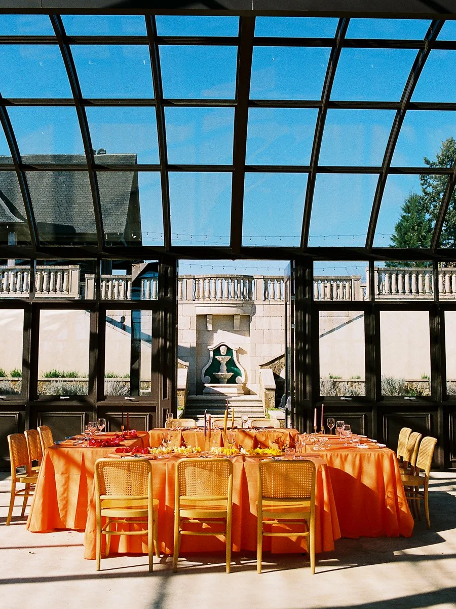 Modern South Asian wedding reception setup with orange tablescapes and cane chairs inside sunlit glass venue.